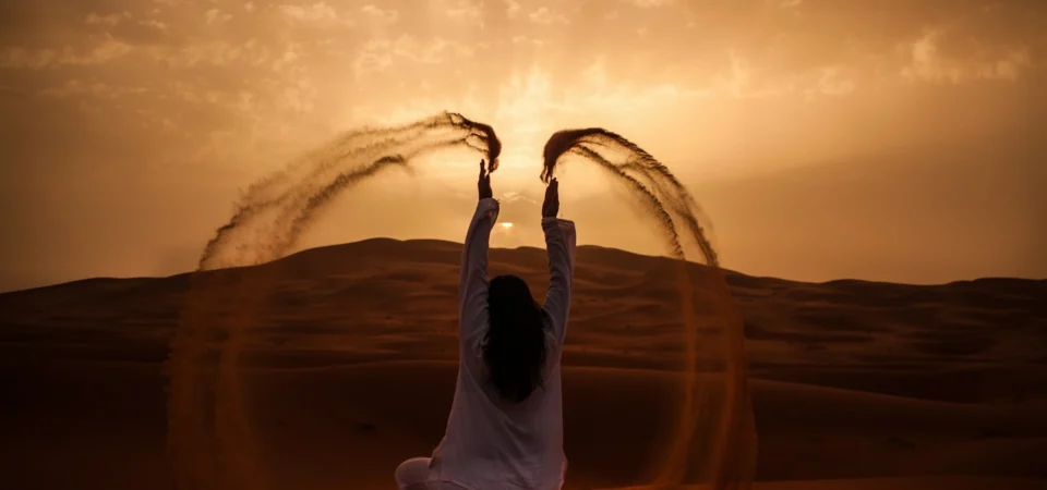A woman dressed in white throws sand into the air while sitting on a dune at sunset in the Moroccan Sahara, creating graceful arcs of golden dust.
