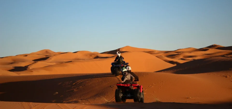 Two travelers on quad bikes ride across the golden sand dunes of Merzouga in the Moroccan desert, kicking up sand under a clear blue sky.