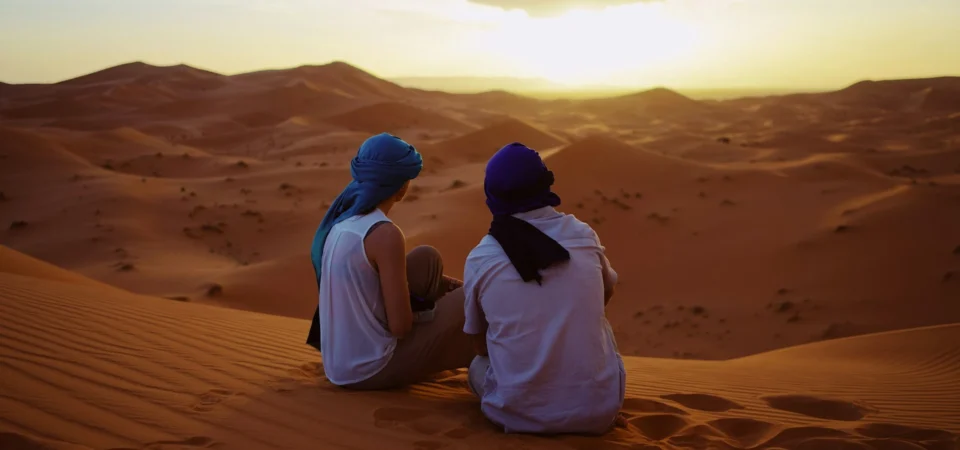 Two travelers wearing blue turbans sit on a sand dune in Merzouga, Morocco, watching the sunset over the rolling desert landscape.
