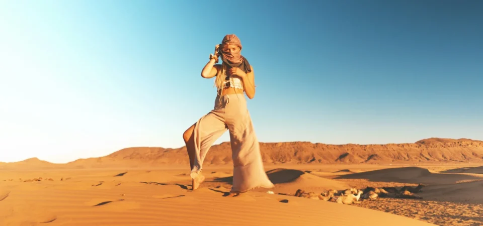 A female traveler poses confidently on the golden dunes of the Moroccan Sahara at midday, wrapped in a desert scarf under a clear blue sky.