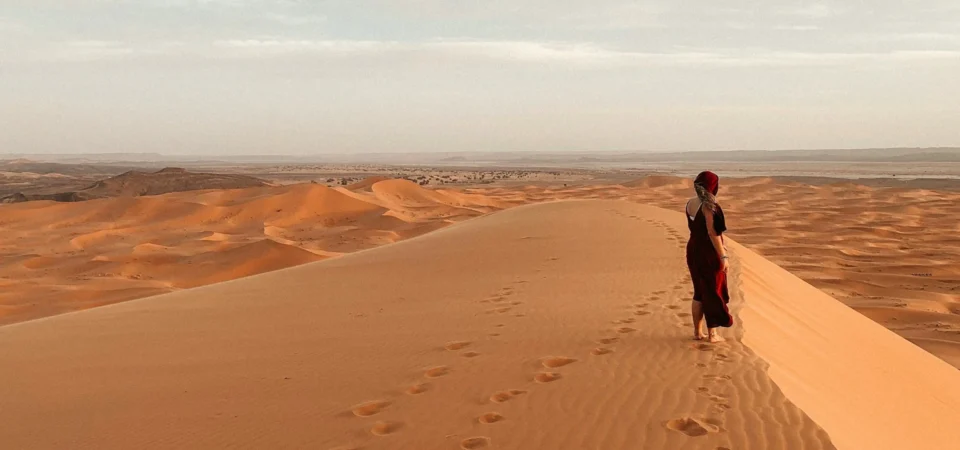 A traveler walks barefoot along a high sand dune in the vast Sahara Desert of Merzouga, Morocco, with golden dunes stretching endlessly into the horizon under a soft, cloudy sky.