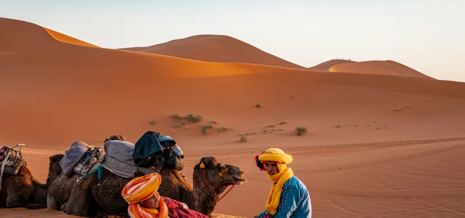 Two Berber men dressed in traditional attire relax beside camels in the Erg Chebbi dunes at sunset in Merzouga, Morocco.